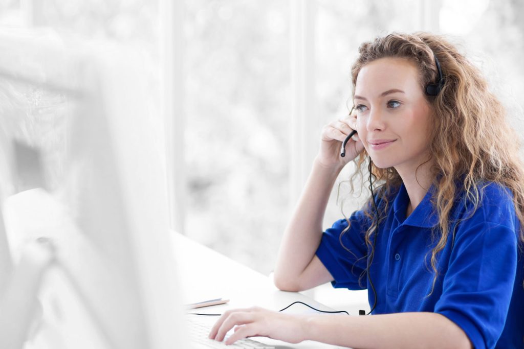 close up smiling friendly female blue shirt operator working with headset in front of computer screen.
